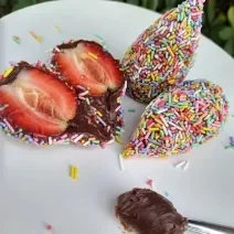 Fotografia em tons coloridos em uma bancada de madeira branca. Ao centro, há um morango cortado ao meio e ao lado, há 2 coxinhas de brigadeiro e uma colher de brigadeiro.