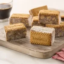 Fotografia de uma tábua de madeira com sete unidades de palha italiana com tostines especiais, ao lado de três biscoitos tostines. No fundo tem uma xícara de vidro com café.