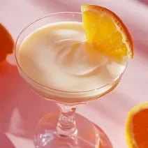 Fotografia de uma taça pequena com uma mousse de laranja e chocolate branco.  Na borda da taça tem um pedaço de laranja, a taça está sobre uma mesa branca com sombras de outras laranjas que estão na mesa.