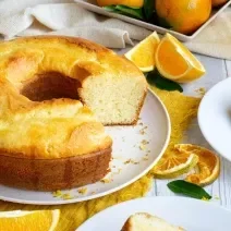 Fotografia de um bolo de laranja feito em forma de furo central sobre um prato raso de cerâmica branco. Ao lado tem um prato de sobremesa com uma fatia do bolo e um garfo, e ao redor tem pedaços e fatias de laranja, sobre uma mesa de madeira branca.