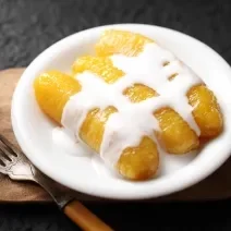 Fotografia de três bananas carameladas com creme de leite por cima.  As bananas estão em um prato pequeno branco raso, ao lado de um garfo que está apoiado em uma tábua de madeira pequena.