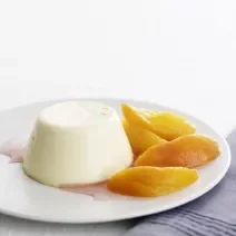 Fotografia em tom claro, com um prato branco sobre uma toalha de mesa branca, ao lado de um pano azul. Dentro do prato, um flan de leite condensado na cor branco, com uma calda clara por baixo, e ao lado, pedaços de pêssego.