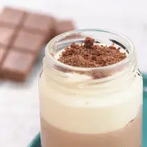 Fotografia em tons de branco e azul de uma bancada branca com um prato retangular azul, sobre ele um copo de vidro com o cheesecake. Ao fundo tabletes de chocolate.