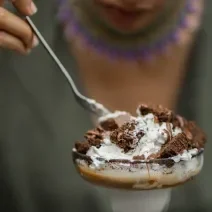 Fotografia de uma pessoa segurando uma colher de sopa e comendo uma sobremesa de camadas de massa e MOÇA Leite Condensado Cremoso. O doce está em uma tacinha de vidro.