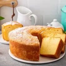 Fotografia de um bolo de laranja feito em forma redonda com furo no meio, o bolo está sobre um apoio, e tem três fatias cortadas, uma apoiada na outra. Ao fundo tem uma fatia do bolo em um prato de vidro pequeno, ao lado de pedaços de laranja.