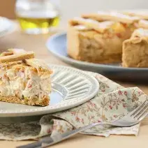 Fotografia em tons de bege em uma mesa de madeira com um pano florido em bege e vermelho e um prato branco apoiado nele com um pedaço de torta de palmito e alho-poró. Ao fundo, um prato grande com a torta cortada ao meio.