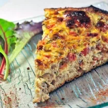 Fotografia mostra uma fatia de torta com camadas bem definidas de massa leve e recheio colorido de sardinha, tomate e azeitonas, polvilhada com orégano.