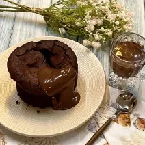 Foto em tons de bege e marrom da receita de bolo fudge servida sobre um prato bege e uma tolha de mesa decorada. Ao lado, há uma colher prateada e uma caneca de vidro com chocolate derretido. Ao fundo, um buqué de flores cor de rosa