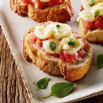 Fotografia em tons de branco e amarelo, com travessa branca e borda dourada com três bruschettas com tomate, mozarela e folhas de manjericão, sobre mesa de madeira rústica.