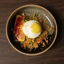 Fotografia de um prato fundo preto de borda marrom, e dentro tem cuscuz, e por cima queijo coalho com calda de café e ovo frito. O prato está sobre uma mesa de madeira.