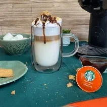 Fotografia em tons de verde em uma bancada com um pano verde, uma xícara de vidro grande com a bebida de leite e café preparada dentro dela. Decorada com marshmallow, calda de chocolate e biscoito triturado.