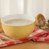 Fotografia em tons de vermelho e amarelo em uma bancada de madeira com um pano xadrez vermelho, uma xícara grande e funda amarela com a bebida de farinha láctea. Ao fundo, um potinho com alguns cereais.