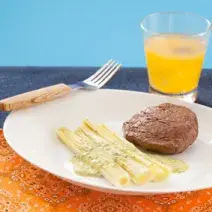 fotografia em tons de branco, azul e laranja de uma bancada vista de frente. Contém um pano laranja, um prato redondo e branco com o alho-poró e um medalhão de filé mignon e ao lado um garfo. Ao fundo um copo transparente com uma bebida laranja