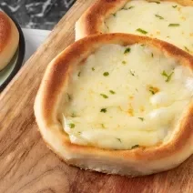 Fotografia de duas esfihas sem glúten de queijo sobre uma tábua de madeira retangular. Ao lado tem um prato em tom de verde com borda preta, e outra esfiha de queijo.