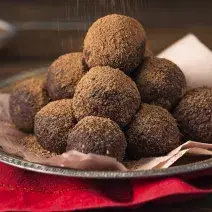 foto em tons de marrom e vermelho de uma bancada marrom vista de frente, contém um pano vermelho, uma bandeja de alumínio com um pano rosa e trufas de chocolate polvilhadas com cacau.