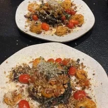 Fotografia de dois pratos de vidro parcialmente quadrados, de pontas arredondadas, sobre uma mesa preta. Dentro dos pratos tem uma massa linguine nero com molho rústico de tomate grape, camarões e farofa de castanhas.