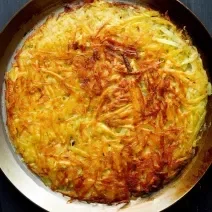 Fotografia em tons de marrom com um prato marrom ao centro. Em cima do prato existe uma receita de batata rosti com batatas raladas e queijo meia cura.