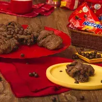 Foto de uma bancada de madeira. Sobre ela há um tecido vermelho, um prato amarelo com um pedaço de pão de Nescau, o pão maior ao fundo sobre um prato vermelho, uma cestinha com pacotes de Nescau Ball e algumas bolinhas de chocolate espalhadas pela mesa.