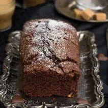Fotografia em tons de marrom de uma bancada marrom vista de cima, contém uma bandeja de alumínio com um bolo inglês de chocolate com açúcar polvilhado por cima.
