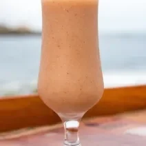 Fotografia de um copo de vidro, estilo taça, com uma bebida de suco de goiaba e neston sobre uma mesa de madeira com vista para a praia.