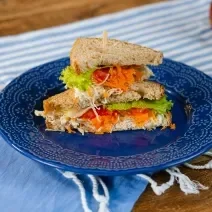 Fotografia em tons de azul com um prato azul ao centro. Em cima do prato existe uma receita de sanduíche natural recheado com frango desfiado, creme de ricota, cenoura, alface e tomate