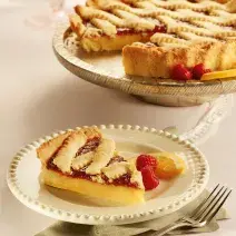 Fotografia em tons de bege em uma mesa de madeira, um guardanapo de pano bege e branco, um prato redondo branco com detalhes bege e uma fatia da crostata com recheio de laranja. Ao fundo, um prato redondo maior com a crostata inteira.