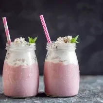 Fotografia de dois recipientes decorativos de vidro com frapê com Leite MOÇA, guaraná e suco de limão. No mesmo copo tem um canudo rosa e branco e hortelã.