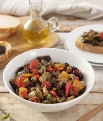 Fotografia de um recipiente branco fundo com caponata de berinjela, ao lado de um prato branco e uma tábua de madeira. 