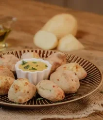 Fotografia de um prato raso escuro com croquetes de batata em volta de um pote branco com molho. 