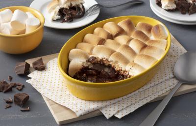 Fotografia em tons de amarelo em uma bancada cinza com um paninho branco de bolinha amarela e um refratário amarelo com o smores de chocolate. Ao fundo, um potinho amarelo com marshmallow e dois pratos com uma fatia do smores em cada um.