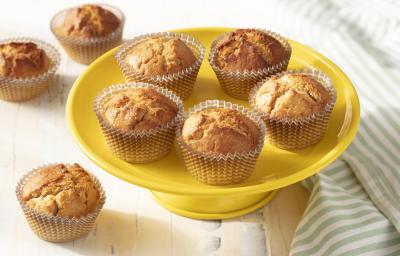 Fotografia em tons de amarelo em uma bancada de madeira branca, um paninho listrado com verde e branco, um prato amarelo alto com vários cupcakes de iogurte com laranja.