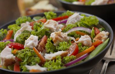 Fotografia em tons de verde, em uma bancada de madeira de cor marrom. Ao centro, um prato redondo preto contendo a salada e ao fundo, um outro prato contendo salada. Ao lado, há um garfo de inox.