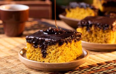 Fotografia em tons de marrom com uma prato pequeno ao centro. Em cima do prato existe uma fatia de um bolo de cenoura de cor alaranjada e com cobertura de chocolate. Ao fundo existe mais fatias desse mesmo bolo.