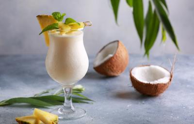 Foto da receita de Piña Colada sem Álcool. Observa-se uma taça grande com a bebida dentro cremosa, uma fatia de abacaxi decorando a borda. Na foto, observa-se um coco seco pela metade.