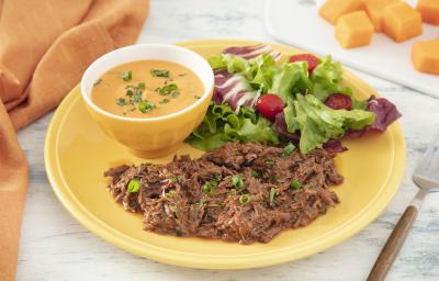 Foto da receita de Purê de Abóbora com Carne Desfiada. Observa-se um prato amarelo com a carne desfiada e a salada e, dentro de um potinho amarelo, o purê de abóbora finalizado com salsinha.