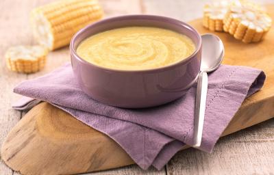 Foto em tons de amarelo da receita de caldinho de milho-verde servida em um bowl de porcelana roxo sobre um paninho roxo também com uma colher prateada ao lado. Tudo sobre uma tábua de madeira e ao fundo espigas de milho cortadas em rodelas