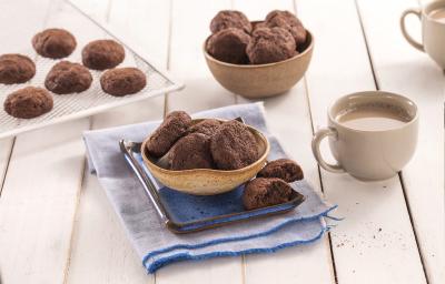 Fotografia em tons de azul em uma bancada de madeira clara, um paninho azul, um potinho bege com alguns cookies de cacau dentro dele. Ao fundo, mais potinhos com cookies e xícaras brancas com café para acompanhar.