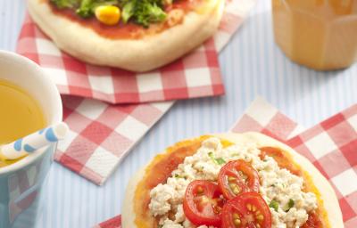 Fotografia em tons de vermelho em uma bancada cinza com guardanapos vermelhos em xadrez e uma mini pizza de legumes e outra mini pizza de tofu apoiadas sobre eles. Ao lado, um copo laranja com suco de laranja e um canudo branco com bolinha azul.