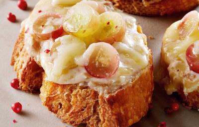 Fotografia em tons de amarelo em uma bancada de madeira escura com um prato branco raso e três bruschettas com queijo brie e uvas.