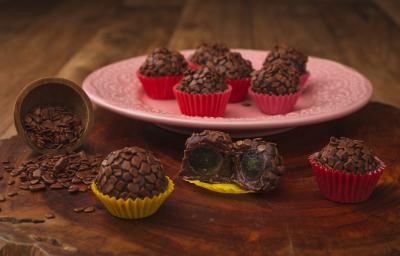 Fotografia em tons de amarelo e vermelho em uma bancada de madeira escura, um prato redondo rosa com brigadeiros em forminhas vermelhas em cima dele. Ao lado, brigadeiros em forminhas amarelas recheados com uva.