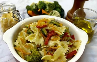 Foto em tons de amarelo da receita de farfalle com legumes servida em uma panela pequena de porcelana vermelha e branca. Ao fundo um vidro de azeite, um triturador de sal, uma porção de legumes e um pote de vidro com farfalle