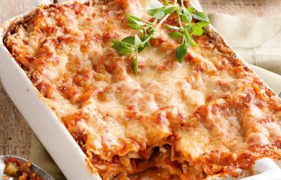 Fotografia em tons de branco e vermelho, ao centro refratário com lasanha de legumes gratinada com queijo, ao lado guardanapo bege com colher de servir, ao fundo prato branco com borda vermelha e porção de lasanha, tudo sobre bancada de madeira rústica.