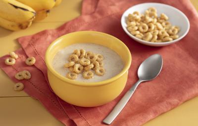 Fotografia em tons de amarelo em uma bancada de madeira amarela com um paninho laranja, um potinho amarelo com o creminho de banana e Cheerios dentro, uma colher e um potinho branco com os cereais Cheerios.