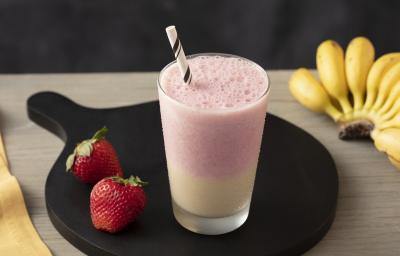 Fotografia em tons claros, em uma bancada de madeira bege escuro e ao lado, um pano amarelo. Ao centro, um prato redondo preto com um copo de smoothie morango e banana e um par de morangos ao lado. Ao fundo, um cacho de bananas.