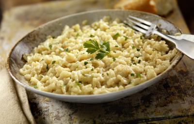 Fotografia de um prato de cor escura com um arroz temperado e salsinha por cima, e ao lado, um garfo apoiado na porta do prato. No fundo da foto, um pão francês cortado na metade, sobre uma toalha de mesa na cor bege.