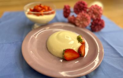 Fotografia em tons de rosa e azul com um prato rosa ao centro. Em cima do prato existe uma receita de Panna Cotta com morangos fatiados por cima