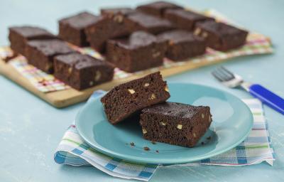 fotografia em tons de azul e marrom de uma bancada vista de frente, contém um pano em tons de branco e azul, um prato azul e redondo contém 2 Brownies e ao fundo um tábua de madeira com diversos Brownies