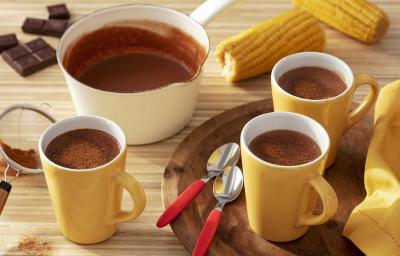 Fotografia em tons de amarelo em uma bancada de madeira de cor marrom. Ao centro, uma bandeja de madeira contendo 2 xícaras amarelas com 2 colheres ao lado. Ao fundo, há 2 espigas de milho, uma panela branca e uma xícara com chocolate quente.