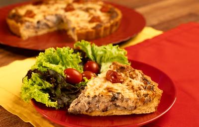 Fotografia mostra um pedaço de quiche cortada sobre um prato vermelho, ao lado folhas verdes de alface e tomate cereja.