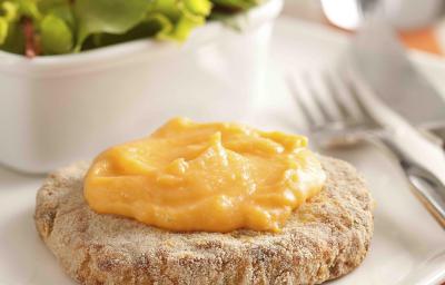 Fotografia em tons de vermelho em uma bancada de madeira com uma toalha listrada vermelha e bege, um prato com o hambúrguer de frango com cheddar derretido em cima. Ao fundo, um potinho com salada de alface.